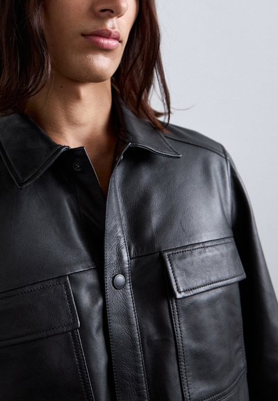 Close-up of a person wearing a black leather jacket with a collar and flap pockets, showing lips and part of the neck and hair.