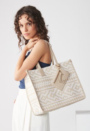 Femeie cu păr creț, purtând un top bleumarin și pantaloni albi, ținând o geantă mare bej cu model și un buzunar cu fermoar atașat.