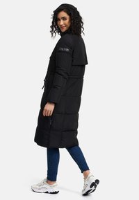 Black puffer coat with a long, loose design, featuring a hood, drawstring waist, and button accents along the side. Styled with dark jeans and sneakers.