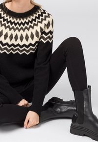 Svart stickad tröja med vita zigzag- och diamantmönster, i kombination med svarta slim-fit jeans och svarta läderanklarboots. Slät textur.