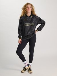 Kvinne med krøllete hår iført svart sportsgenser med lange ermer, svarte leggings, hvite stripete sokker og gullfargede sportssko, står med hånden på hoften.