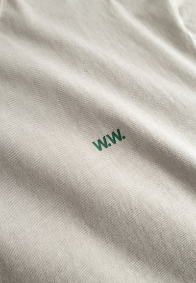 Gros plan sur un tissu beige clair avec un petit texte « W.W. » vert imprimé à la surface.