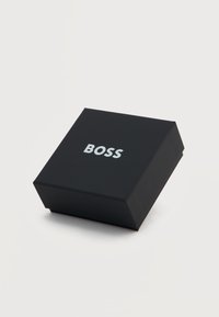 Μαύρο τετράγωνο κουτί με τη λέξη "BOSS" τυπωμένη με άσπρα γράμματα στην κορυφή, τοποθετημένο σε μια απλή ανοιχτόγριζη επιφάνεια.