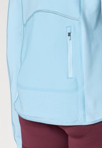 Top atlético azul-claro com um padrão texturizado, bolso lateral com fecho, e costuras contrastantes, combinado com leggings bordô.