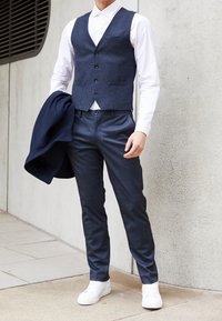 Hombre con chaleco azul oscuro, pantalones de cuadros en tonos navy, camisa blanca y zapatillas blancas sosteniendo un abrigo navy contra un fondo de pared de concreto.
