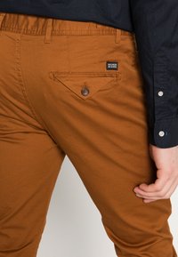 Braune Chinos aus Baumwolle, mit einer Gesäßtasche mit Knopfverschluss und einem kleinen Logo-Tag. Glatte Textur und taillierte Passform.