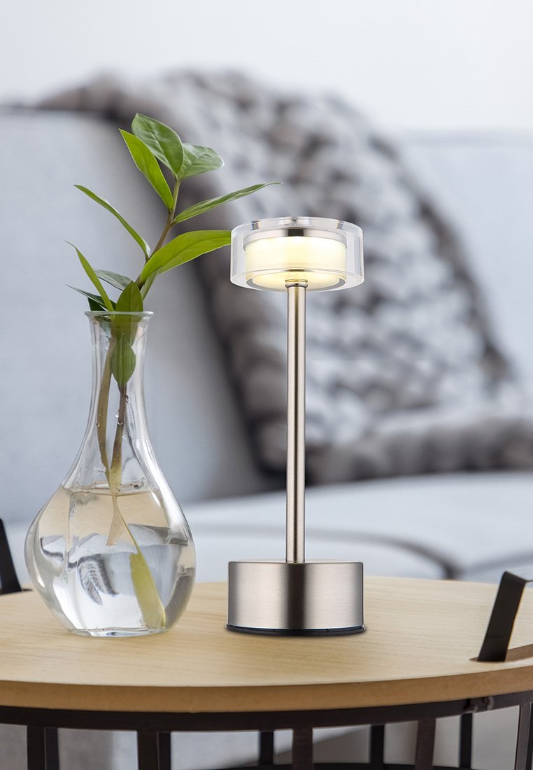 Lampa stołowa z przezroczystym kloszem, metalicznie srebrną podstawą, cylindrycznym kształtem i miękkim białym światłem; ustawiona obok szklanej wazy z rośliną.