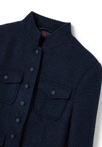 Giacca strutturata blu navy con colletto alla mandarino, cinque bottoni e due tasche sul petto con chiusura a patta. Il materiale è tessuto intrecciato.