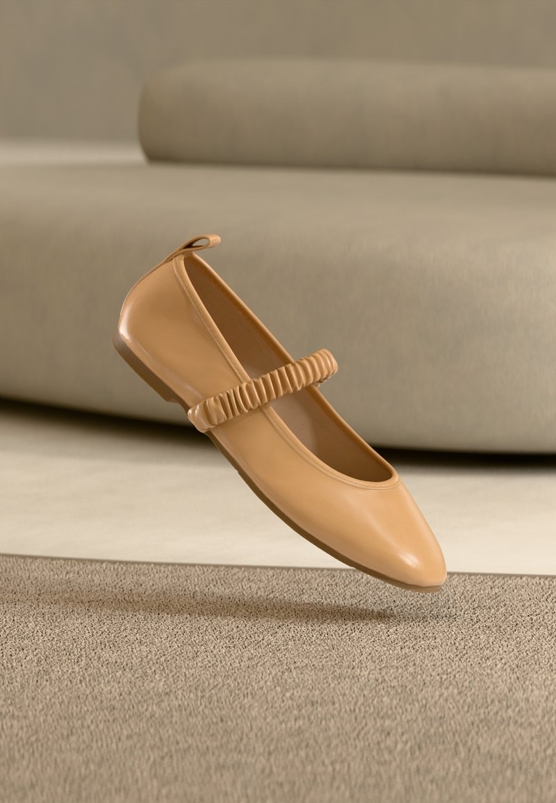 Chaussure Mary Jane plate marron clair avec bride froncée flottant au-dessus d'un tapis beige devant un canapé assorti.