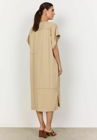 Knielange, beige jurk met korte mouwen, zijsplitten en zichtbare stikdetails. Gemaakt van een lichte stof met een comfortabele pasvorm.