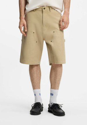 Mann trägt beige knielange Shorts mit Vordertaschen, weiße Socken mit blauem Logo und schwarze Schnür-Loafer, steht vor weißem Hintergrund.