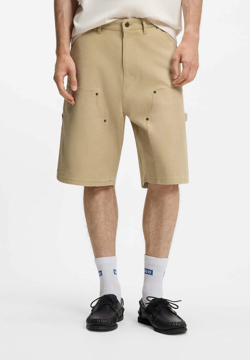 Homme portant un short beige longueur genou avec poches avant, chaussettes blanches avec logo bleu et mocassins noirs à lacets, debout sur fond blanc.