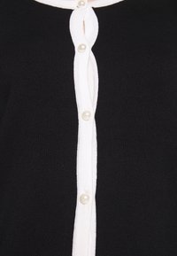 Svart stickad cardigan med en vit knappslå och pärlknappar. Slät textur och en klassisk design med en lätt V-ringning.