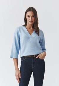 Lyseblå sweater med V-udskæring og trekvartærmer, lavet af blødt materiale. Sammen med mørkeblå jeans og et brunt bælte.