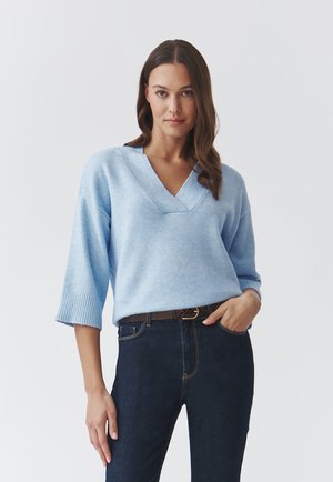 Hellblauer Pullover mit V-Ausschnitt und Dreiviertelärmel, aus weichem Material. Kombiniert mit dunkelblauen Jeans und einem braunen Gürtel.