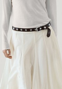 Zwarte leren riem met zilveren studs, gedragen over een witte, luchtige, geplooide rok. Langer mouwen, lichtgekleurde top en een ontspannen handpositie zichtbaar.