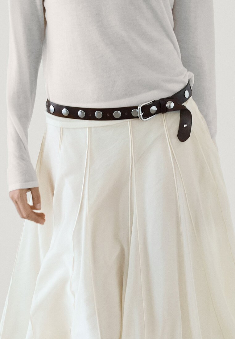 Zwarte leren riem met zilveren studs, gedragen over een witte, luchtige, geplooide rok. Langer mouwen, lichtgekleurde top en een ontspannen handpositie zichtbaar.