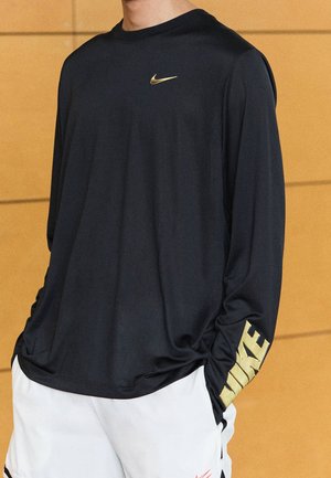 Persona vistiendo una camiseta negra de manga larga de Nike con logo dorado en el pecho y la manga, combinada con pantalones cortos blancos, frente a un fondo de pared de madera.