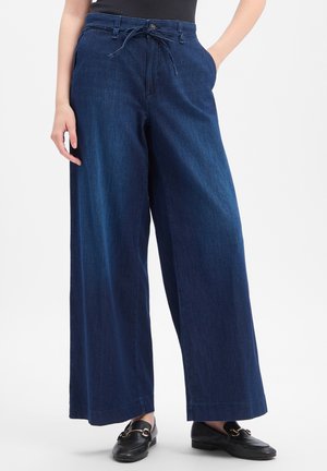 Frau trägt dunkelblaue weit geschnittene Jeans mit Kordelzug in der Taille, schwarzes Hemd und schwarze Loafer, steht mit den Händen in den Taschen.