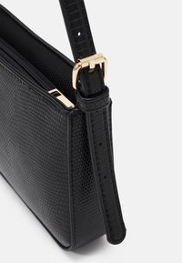 Sort tekstureret læderhåndtaske med lynlås, udstyret med en guldfarvet spænde på en justerbar rem og en struktureret form.