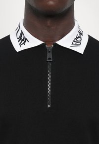 Svart glidelås polo skjorte med strukturert stoff og hvit krage med trykt merkevare. Glidelåsen er plassert midt foran.