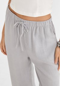 Nainen, joka pitää yllään vaaleanharmaita joustavia vyötärönyöripantseja, joissa on sivutaskut, ja valkoista keskipitkää toppia, seisoo kädet sivuilla.