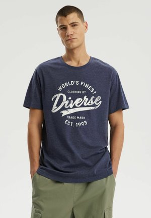 Młody mężczyzna stoi z rękami w kieszeniach, ubrany w granatowy t-shirt z logo "Diverse" i oliwkowe spodnie typu cargo na jednolitym tle.