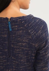 Modelka od tyłu w granatowym swetrze o fakturze z widocznym zamkiem błyskawicznym. Materiał ma tkany wzór z jaśniejszymi akcentami.