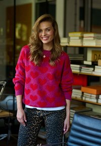 Chlupatý růžový svetr s červenými srdíčkovými vzory, doplněný o kalhoty s leopardím potiskem. Prostředí obsahuje knihovny a moderní nábytek.