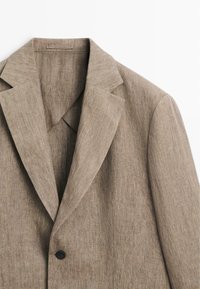 Beige linnen blazer met inkepingrevers en een enkele zwarte knoopsluiting, getoond tegen een witte achtergrond.
