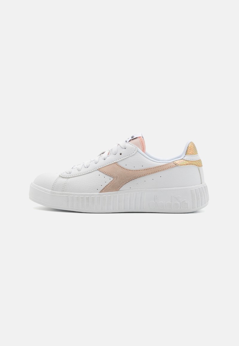 Zapatilla baja blanca con logo beige en el lateral, acento rosa en la lengüeta y detalle de talón texturizado en dorado sobre fondo blanco.