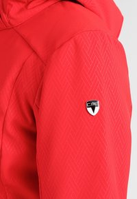 Rød isoleret jakke med fiskebensmønster og et logo-patch på ærmet. Stoffet fremstår glat og holdbart.