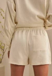 Cremfarbene Shorts aus strukturiertem Stoff, mit elastischem Bund, Seitentasche und rohem Saum. Einfaches, lässiges Design.