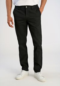 Hombre con pantalones rectos negros y zapatillas blancas, de pie con una mano en el bolsillo y la otra relajada a su lado.