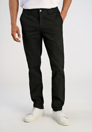 Hombre con pantalones rectos negros y zapatillas blancas, de pie con una mano en el bolsillo y la otra relajada a su lado.