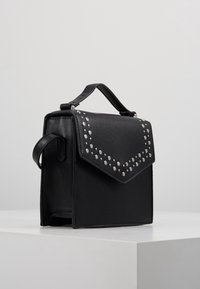 Czarna torebka ze sztucznej skóry o prostokątnym kształcie, z klapą zapinaną na magnes, ozdobioną srebrnymi ćwiekami wzdłuż krawędzi, oraz uchwytem u góry.
