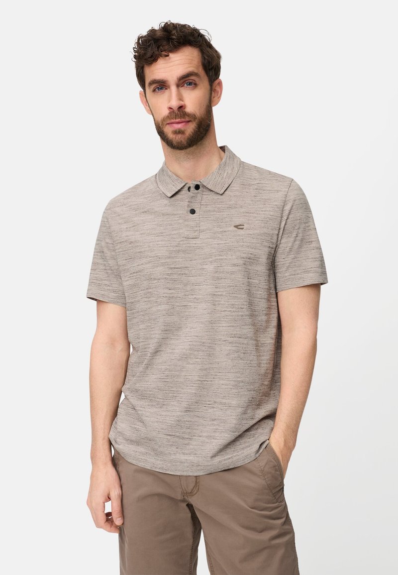 Mand med skæg iført beige kortærmet polo med to sorte knapper og brune bukser, stående med den ene hånd i lommen.