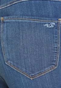 Blå denimjeans bakficka med kontrasterande orange sömnad och en liten vit broderad måslogotyp nära det övre högra hörnet.