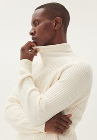 Baltas raukšlėtas golfas su prigludusia siluetu ir tekstūruotu mezgimu. Modelis pateiktas šone, su rankomis arti apykaklės.