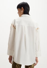 Camicia bianca a maniche lunghe con spalle scoperte, dettagli con cravatta sulle maniche e orlo dritto. Tessuto morbido e colletto button-down.
