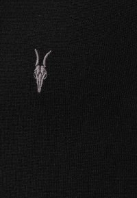 Tejido de punto negro con un pequeño diseño bordado de un cráneo de cabra gris en el lado izquierdo, con una textura fina y un acabado suave.