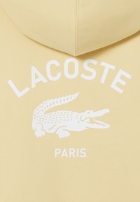 Sudadera amarilla con un logo de cocodrilo impreso en blanco y el texto "LACOSTE PARIS". Fabricada en tejido suave con un corte relajado.