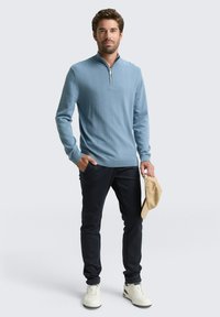 Hellblaues, mit Reißverschluss versehenes Sweatshirt mit einem strukturierten Muster, kombiniert mit dunklen Hosen und weißen Turnschuhen. Das Modell hält eine beigefarbene Mütze.