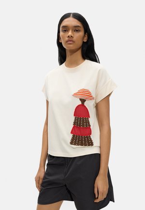 Kobieta ubrana w kremowy top z krótkim rękawem z koralikowym czerwono-brązowym wzorem grzyba, połączony z czarnymi szortami na jednolitym tle.