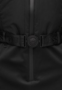 Casaco preto com fecho central e cinto ajustável preto com fivela redonda gravada com um padrão floral e o texto "GUESS".