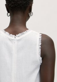 Weißes ärmelloses Oberteil mit fransigen Kanten, aus einem strukturierten Stoff gefertigt. Verfügt über einen unauffälligen Reißverschluss auf der Rückseite und eine schlichte, klare Silhouette.