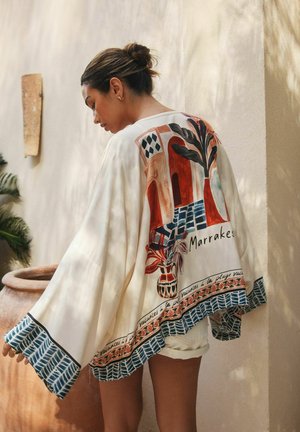 Frau mit hochgestecktem Haar trägt einen lockeren cremefarbenen Kimono mit buntem Marrakesch-Motiv und blau gemustertem Saum, sie steht an einer beigefarbenen Wand.