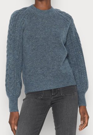 Personne portant un pull bleu en maille torsadée avec des manches ballon et un jean gris foncé ajusté, debout devant un fond clair.
