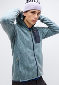 Νέος άνδρας που ρυθμίζει τη κουκούλα ενός ανοιχτό μπλε φλις μπουφάν με σχέδια και σκούρο μπλε τσέπη στο στήθος, φοράει ένα μωβ σκουφάκι με μαύρο κείμενο.