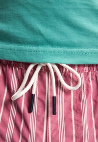 Maglietta in cotone turchese con orlo sopra i costumi da bagno a righe rosa con cordino bianco, dotato di punte in plastica nere.